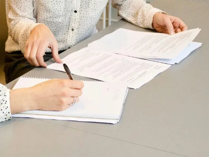 Manos firmando y revisando documentación sobre una mesa. Sin rostros ni datos personales legibles.