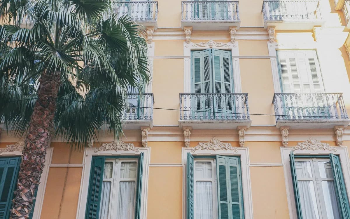 Fachada de un edificio de viviendas español con balcones de forja, contraventanas verdes y una palmera delante.