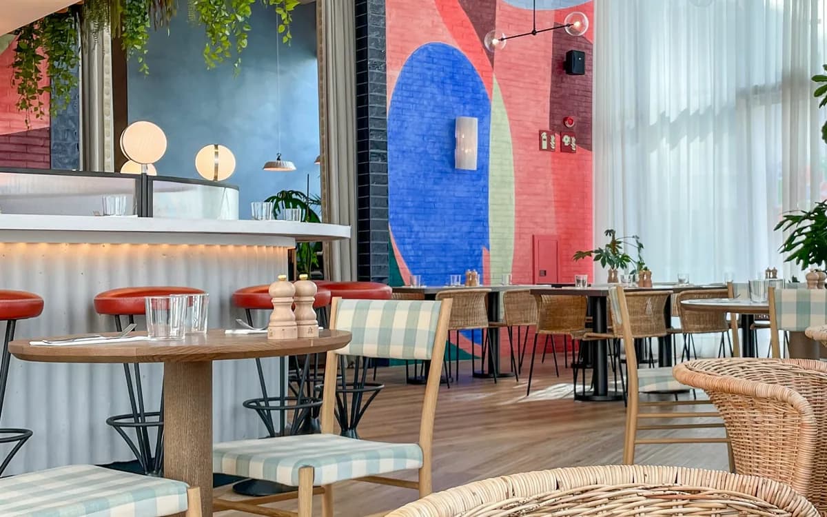 Comedor de un restaurante con barra de taburetes rojos, mesas de madera y sillas de mimbre, iluminado por ventanales.