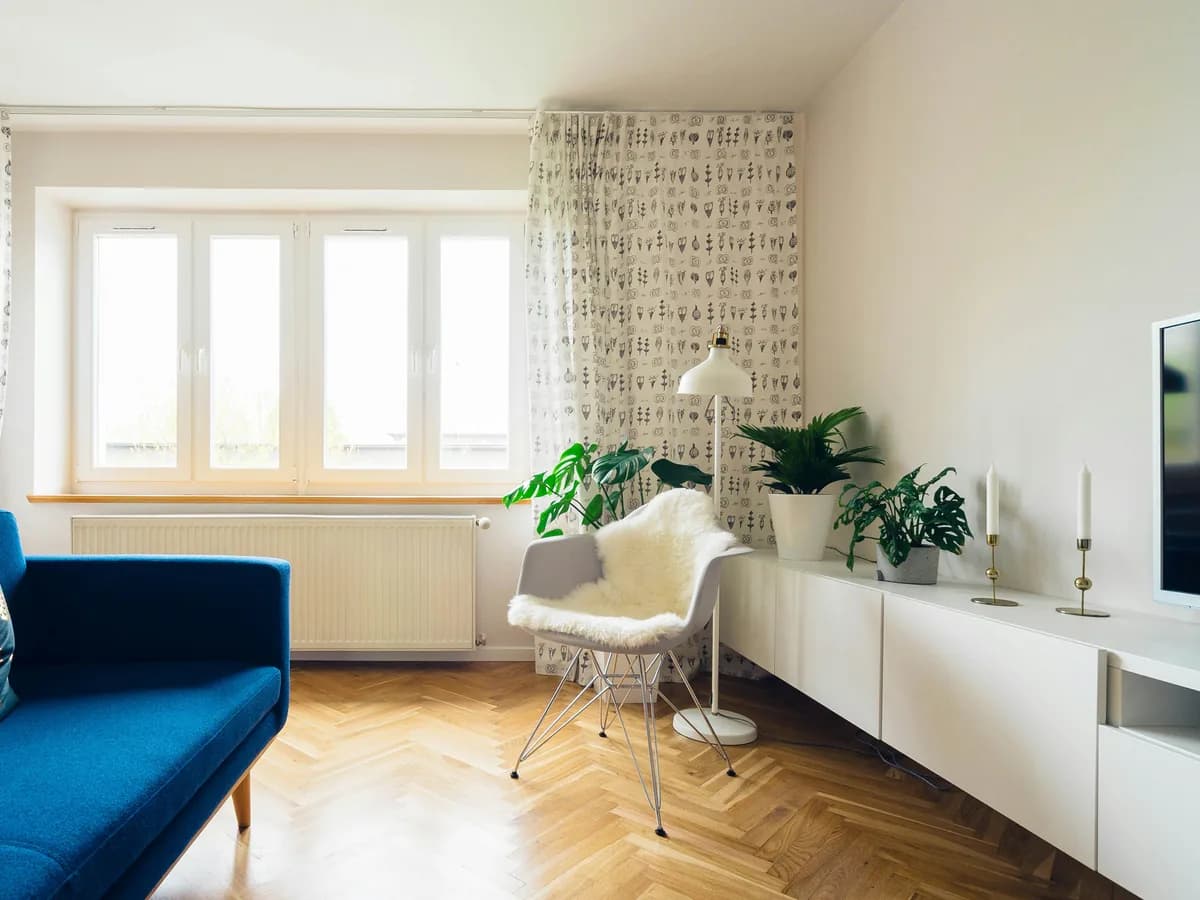 Salón de una vivienda con luz natural: suelo de madera en espiga, sofá azul, butaca junto a la ventana y plantas sobre un aparador.