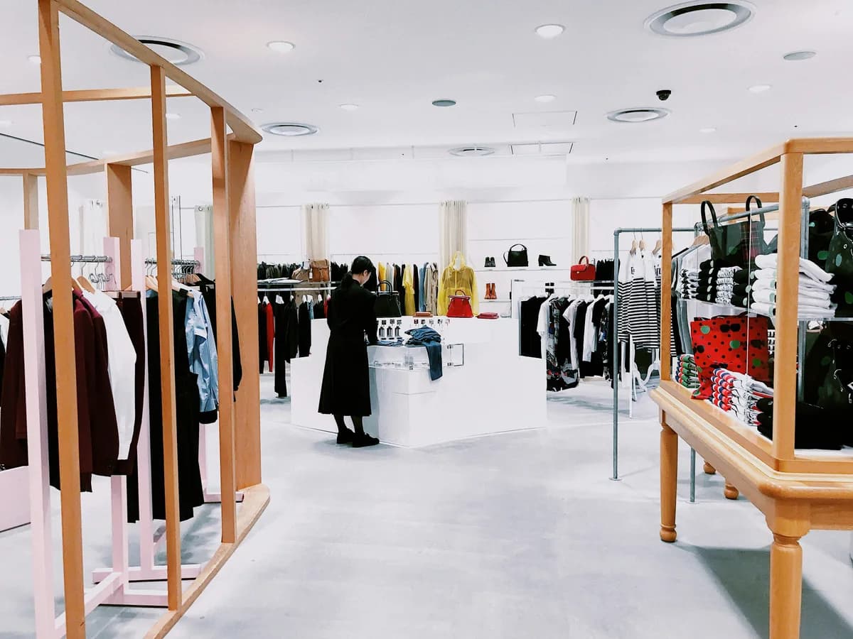 Interior de una tienda de ropa con percheros, expositores de madera clara y una clienta de espaldas mirando un mostrador.